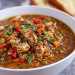 A hearty bowl of chicken lentil soup filled with tender chicken pieces, lentils, carrots, and fresh herbs, served with crusty bread on the side.