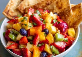 A colorful bowl of fresh fruit salsa made with strawberries, kiwi, and pineapple, served alongside golden cinnamon sugar tortilla chips on a wooden platter.