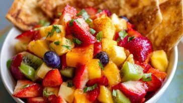 A colorful bowl of fresh fruit salsa made with strawberries, kiwi, and pineapple, served alongside golden cinnamon sugar tortilla chips on a wooden platter.