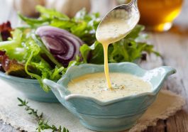A glass jar of homemade parmesan vinaigrette with visible parmesan shreds, surrounded by fresh greens and lemon slices.