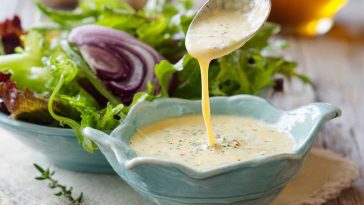 A glass jar of homemade parmesan vinaigrette with visible parmesan shreds, surrounded by fresh greens and lemon slices.