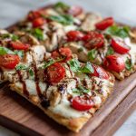 A golden flatbread topped with grilled chicken, melted mozzarella, fresh tomatoes, basil leaves, and a drizzle of balsamic glaze, served on a wooden board.