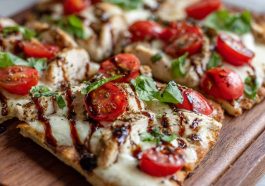 A golden flatbread topped with grilled chicken, melted mozzarella, fresh tomatoes, basil leaves, and a drizzle of balsamic glaze, served on a wooden board.