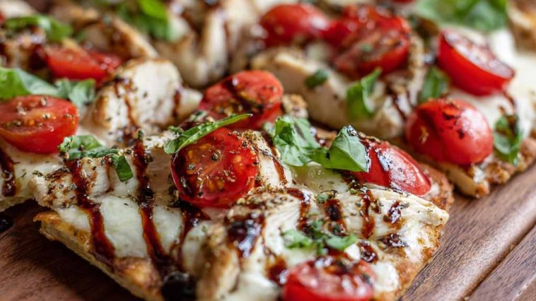 A golden flatbread topped with grilled chicken, melted mozzarella, fresh tomatoes, basil leaves, and a drizzle of balsamic glaze, served on a wooden board.