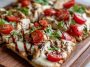 A golden flatbread topped with grilled chicken, melted mozzarella, fresh tomatoes, basil leaves, and a drizzle of balsamic glaze, served on a wooden board.