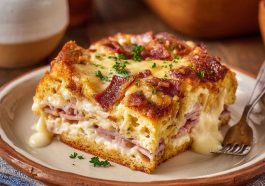 A golden-baked Monte Cristo breakfast casserole layered with ham, turkey, Swiss cheese, and custard, topped with powdered sugar and served with raspberry jam.