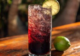A tall glass of Midnight Malibu Dirty Soda layered with cola, coconut syrup, and lime, topped with ice cubes and a lime wedge garnish.