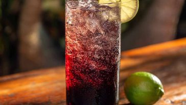 A tall glass of Midnight Malibu Dirty Soda layered with cola, coconut syrup, and lime, topped with ice cubes and a lime wedge garnish.