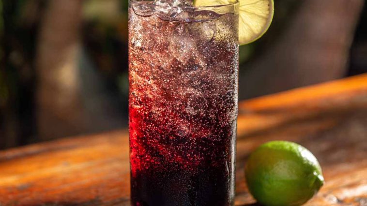 A tall glass of Midnight Malibu Dirty Soda layered with cola, coconut syrup, and lime, topped with ice cubes and a lime wedge garnish.