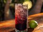 A tall glass of Midnight Malibu Dirty Soda layered with cola, coconut syrup, and lime, topped with ice cubes and a lime wedge garnish.