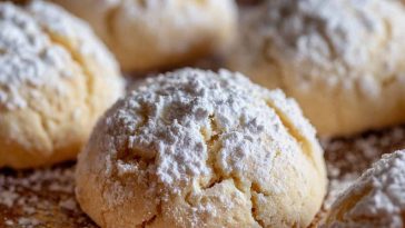 Golden pizzicati Italian pinch cookies filled with jam, lightly dusted with powdered sugar, arranged on a rustic wooden board.