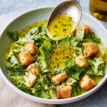 A small glass jar of homemade parmesan vinaigrette dressing with fresh salad greens in the background.