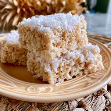 Coconut Rice Krispy treats topped with toasted coconut flakes and cut into squares on a dessert tray.