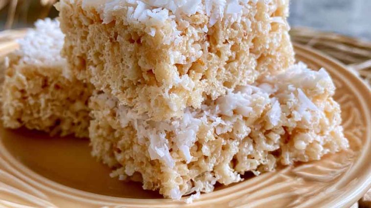 Coconut Rice Krispy treats topped with toasted coconut flakes and cut into squares on a dessert tray.